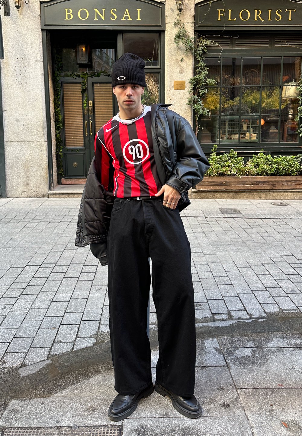 Maillot rayé rouge et noir avec logo circulaire, veste noire surdimensionnée, pantalon noir à jambes larges et bottes noires, sur fond de ville.