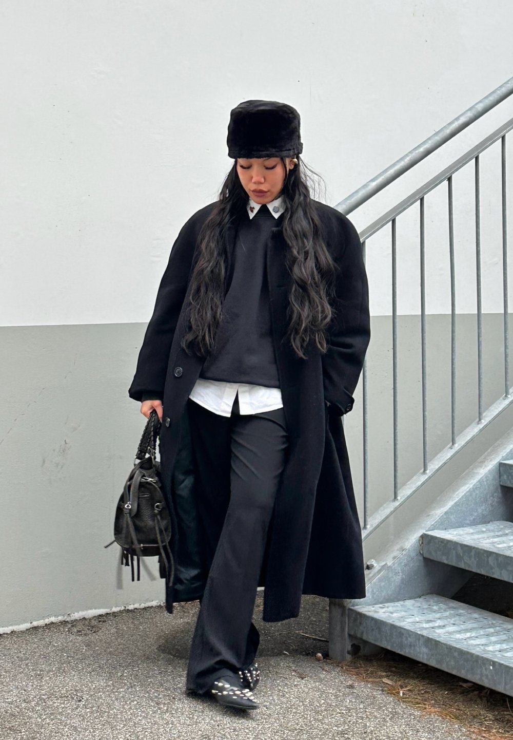Lange zwarte jas over een zwarte trui en wit shirt; zwarte broek; zwarte (nep)bonten hoed; draagt een kleine zwarte tas; gestructureerde schoenen met studs.