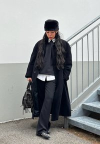 Lange zwarte jas over een zwarte trui en wit shirt; zwarte broek; zwarte (nep)bonten hoed; draagt een kleine zwarte tas; gestructureerde schoenen met studs.