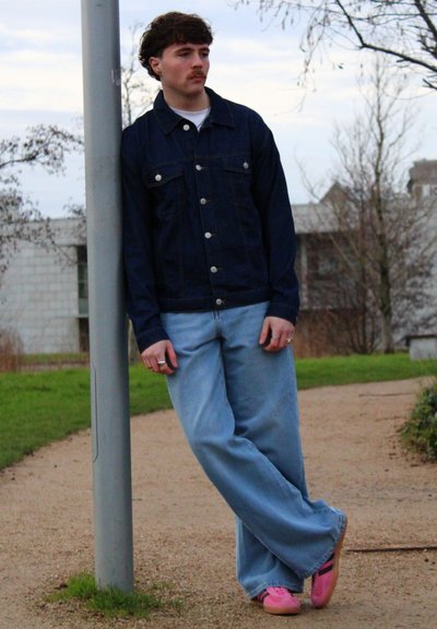 Hombre con bigote vestido con una chaqueta de mezclilla oscura, jeans azul claros y zapatos rosas, apoyado en un farola sobre un camino de grava al aire libre.