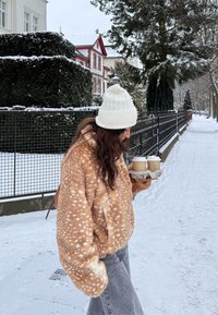 Femme portant un bonnet en tricot blanc et une veste en fourrure marron à taches, marchant sur un trottoir enneigé, tenant deux gobelets de café dans un porte-gobelets en carton.
