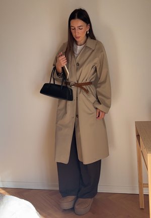 Abrigo trench beige con grandes botones, cintura ceñida con cinturón y mangas oversize. Combinado con pantalones oscuros y bolso negro. Calzado suave y texturizado.