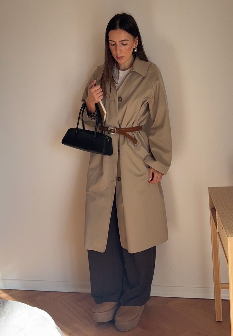 Beige trench coat with large buttons, belted waist, and oversized sleeves. Paired with dark trousers and black handbag. Soft, textured footwear.