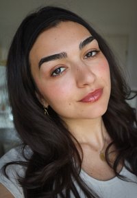 Vrouw met lang, donker haar in een witte top. Gouden oorbellen en een gouden ketting zichtbaar. Een heldere huid met een natuurlijke make-up look.