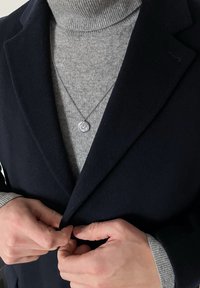 Personne boutonnant un manteau bleu marine foncé par-dessus un pull à col roulé gris clair, portant un collier pendentif argenté avec un motif gravé.