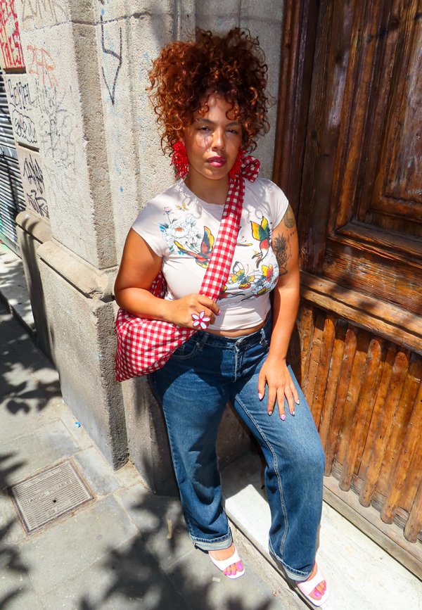 Jeune femme aux cheveux bouclés se tient contre un mur couvert de graffitis et une porte en bois, portant un haut fleuri, un jean et un sac à carreaux rouge.