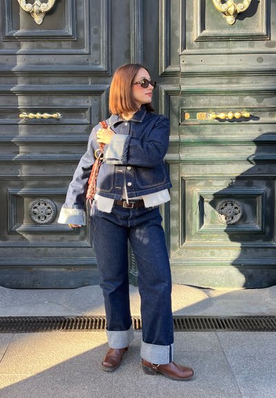 Chaqueta de mezclilla y pantalones con puños de color azul claro, llevados con un cinturón marrón. Gafas de sol con montura negra y botas de tobillo marrones. Fondo de puerta verde.