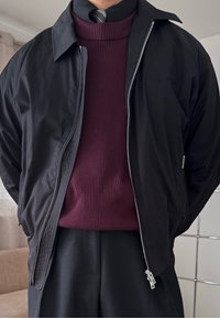 Άτομο που φοράει μαύρο σακάκι πάνω από πουλόβερ μπορντό με ριγέ πλέξη και μαύρο πουκάμισο με γιακά, με γκρι ριγέ γραβάτα.