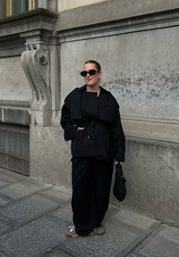 Kobieta w czarnym płaszczu, szerokich czarnych spodniach, okularach przeciwsłonecznych i sneakersach, stojąca przy kamiennym budynku, trzymająca w ręku fakturowaną czarną torebkę.