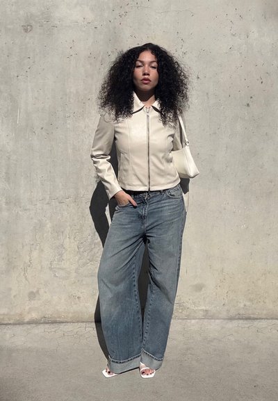 Mujer con pelo rizado que lleva una chaqueta beige con cremallera, vaqueros de pata ancha, tacones blancos abiertos y lleva un bolso blanco de hombro, de pie junto a una pared de hormigón.