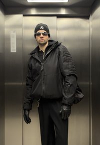 Homme portant une veste noire, des gants, un bonnet et des lunettes de soleil, debout à l'intérieur d'un ascenseur métallique avec un sac à dos sur une épaule.