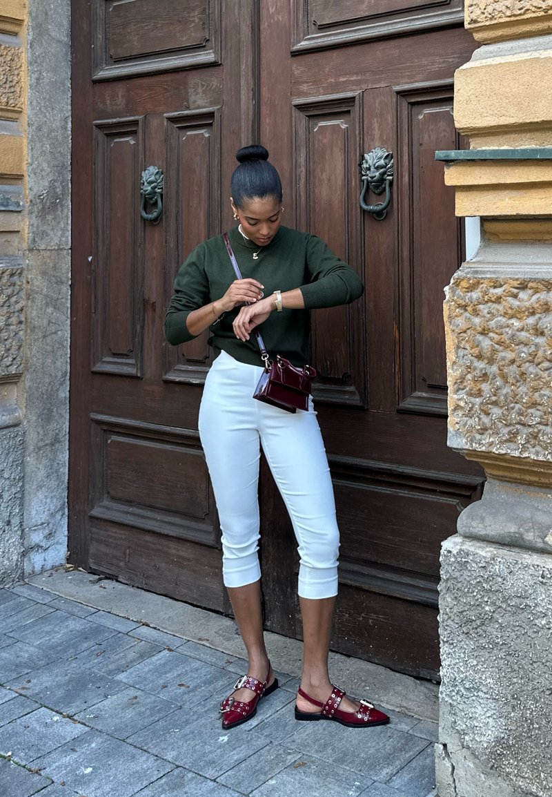 A woman wearing white cropped trousers, a dark green sweater, and burgundy shoes and handbag is standing on stone paving, checking her wristwatch in front of a large wooden door.