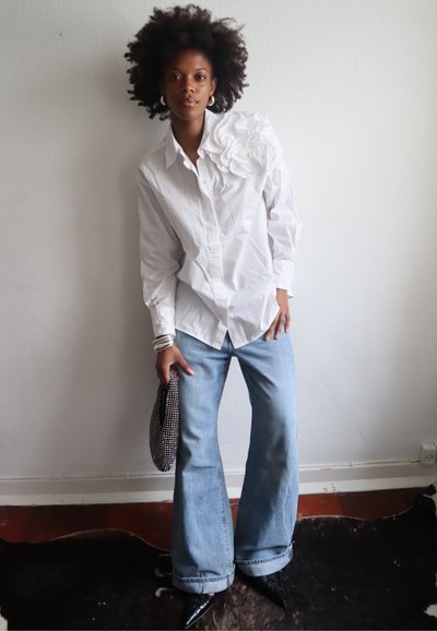 Camisa blanca de botones con un aplique floral en el hombro izquierdo, combinada con jeans acampanados de lavado claro y un bolso de mano con estampado en blanco y negro.