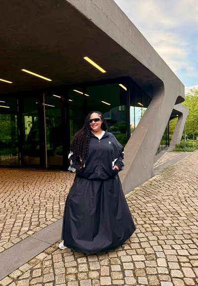 Chaqueta negra con acentos blancos y una falda negra, con un fondo de un edificio moderno de diseño geométrico. Sendero de piedra pavimentado y vegetación visible.