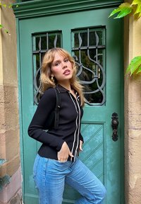 Cardigan a coste nera con bordi bianchi, abbinato a jeans azzurri chiari. In piedi accanto a una porta verde con maniglie decorative nere.