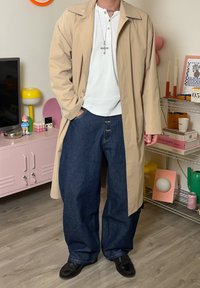 Bēšs viegls trančs mētelis pār baltu Henley krekliņu, kopā ar ziliem platu kāju džinsiem ar izteiktām pogām un melniem apaviem.