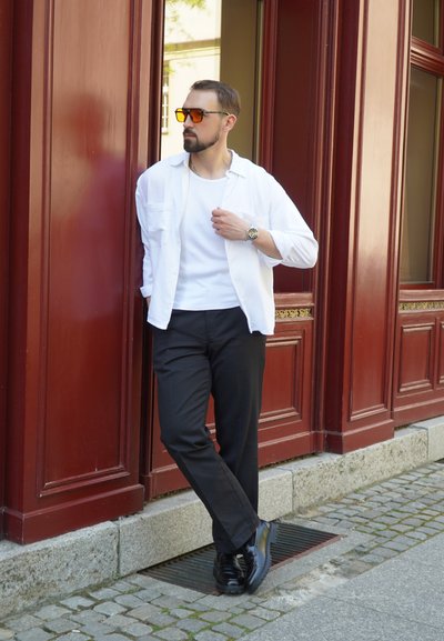 Camisa blanca de botones sobre una camiseta blanca sin mangas, pantalones negros y zapatos negros. El fondo presenta paneles de madera rojos y pavimento de piedra.