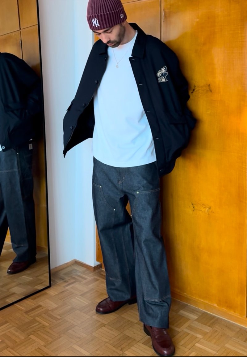 Veste noire avec détails graphiques, t-shirt blanc, pantalon en denim surdimensionné et chaussures marron, debout à côté d'un miroir contre un panneau en bois.