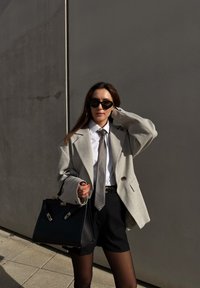 Grijze oversized blazer over een wit shirt, zilverkleurige stropdas, zwarte korte broek en doorschijnende panty's. Zwarte handtas met gouden accenten, zonnebril.