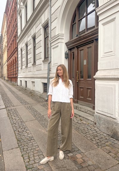 Mujer con cabello largo rubio, vestida con una camisa blanca y pantalones amplios de olivo, de pie en una acera de adoquines junto a un edificio blanco con una gran puerta de madera.