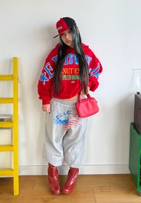 Sweat à capuche rouge oversize avec texte bleu et jaune, pantalon de survêtement gris avec imprimé bleu et rouge, bottines rouges brillantes et petit sac à main rouge.