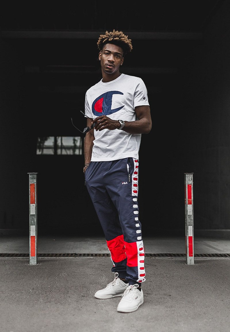 White t-shirt with a blue and red logo, teamed with navy track pants that have red accents and white side details. The outfit is completed with white sneakers.