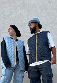Two individuals are wearing quilted vests; one is light blue with a front zip and dark accents, while the other is dark blue with a multicoloured zipper.