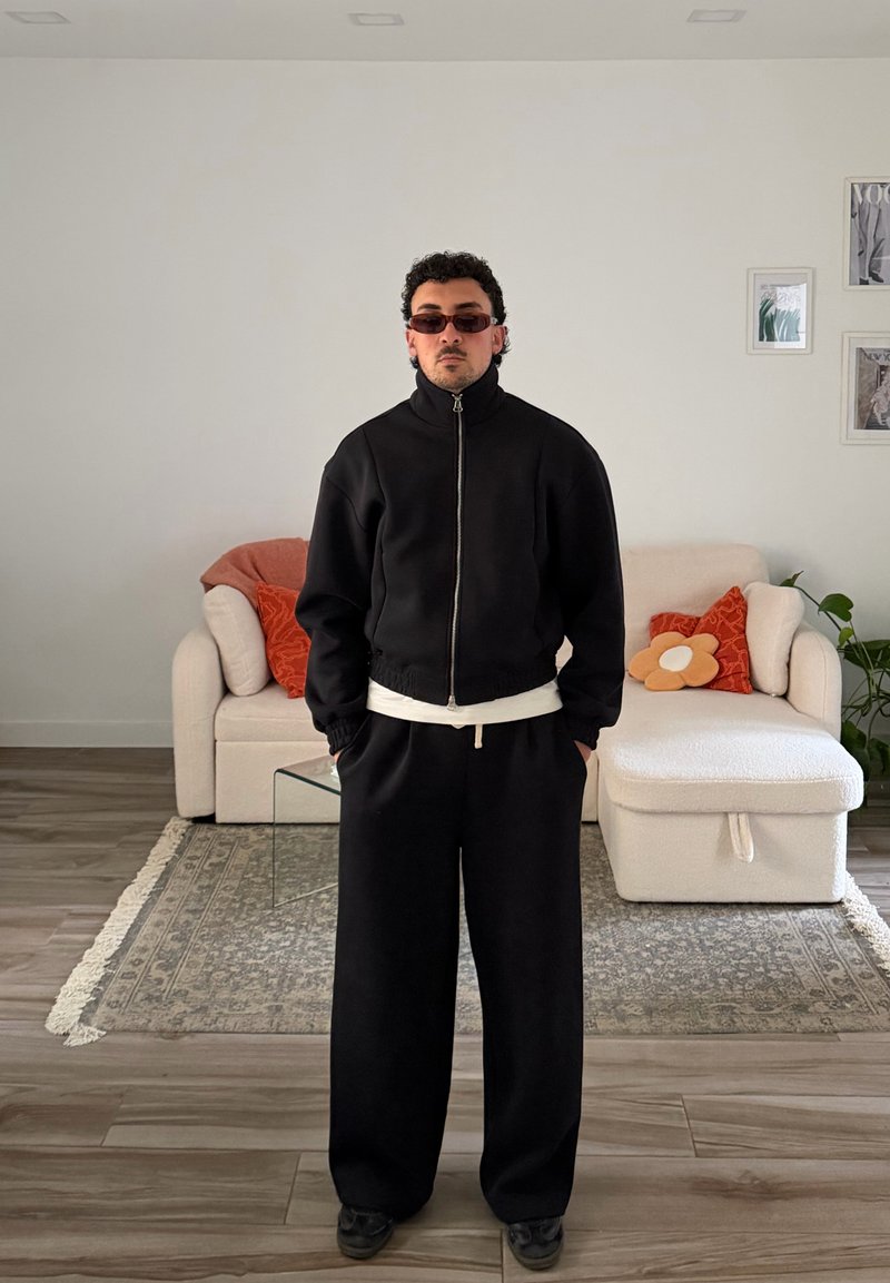 Homem com cabelo curto e encaracolado e óculos de sol está em pé dentro de casa, usando um casaco preto com fecho éclair e calças largas, com um sofá branco e almofadas laranjas atrás dele.