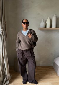 Γυναίκα με σκουρόχρωμα γυαλιά ηλίου, καφέ πουλόβερ με V λαιμόκοψη, παντελόνι φαρδύ και κρατώντας καφέ τσάντα σε δωμάτιο με ουδέτερους τόνους και κεραμικά βάζα.