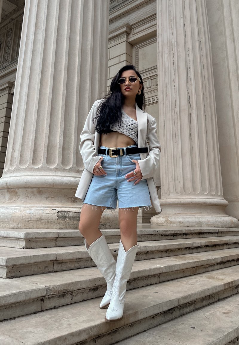 Λευκό προσαρμοσμένο σακάκι πάνω από πλεκτό crop top, ανοιχτό παντελόνι τζιν με φθαρμένες άκρες, και λευκές καουμπόικες μπότες. Τα αξεσουάρ περιλαμβάνουν γυαλιά ηλίου και μια μαύρη ζώνη.