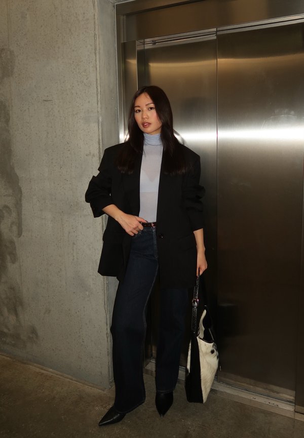 Sieviete ar gariem melniem matiem, valkājot melnu žaketi, gaiši zilu augstu apkaklīti, tumšas džinsas un melnus smailus apavus, stāv pie metāla lifta.