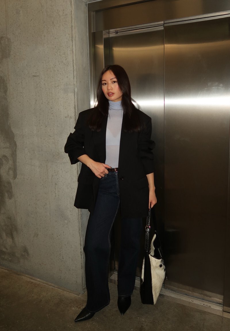 Femme aux longs cheveux noirs portant un blazer noir, un col roulé bleu clair, un jean foncé et des chaussures noires à bout pointu, debout près d'un ascenseur métallique.