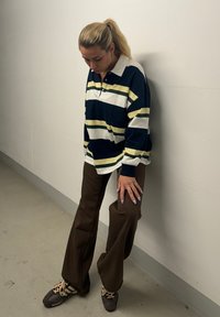 Langarm-Polohemd in Marineblau und Weiß mit gelben Akzenten, kombiniert mit braunen Schlaghosen und braunen Sneakers mit cremefarbenen Details.