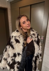 Črno-bela kosmata bunda, prevrnjena čez ramena, pod njo temen pulover. Tekstura je puhasta z rahlim sijajem.