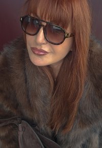 Frau mit langen roten Haaren, die eine übergroße Schildpatt-Sonnenbrille und einen braunen Pelzmantel trägt, während sie eine dunkelbraune Wildledertasche hält.
