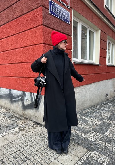 Mujer con un abrigo negro, gorro rojo, gafas de sol y pantalones a rayas está de pie en una acera de baldosas cerca de la esquina de un edificio rojo con ventanas.
