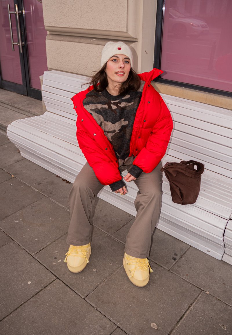 Veste matelassée rouge, pull camouflage, pantalon cargo beige et chaussures à lacets jaunes. Un sac marron repose sur un banc blanc dans un cadre urbain.