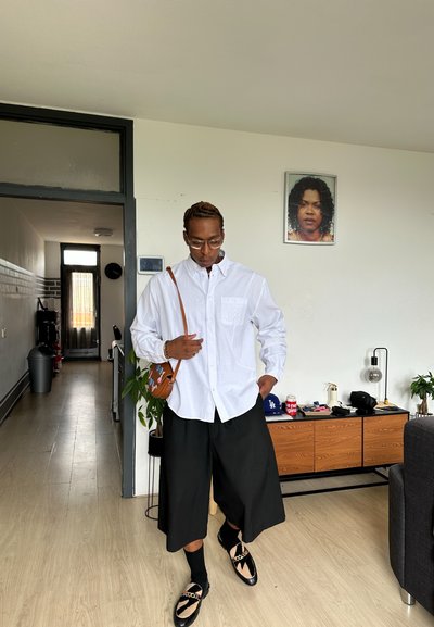 Camisa blanca de botones con un bolsillo, pantalones cortos anchos negros y calzado negro con un patrón. Bolso marrón cruzado con un acento. Ambiente casual en interiores.