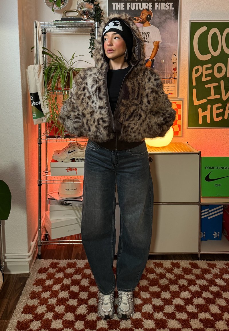 Veste courte en fausse fourrure à imprimé léopard avec capuche, ceinture côtelée foncée et manches longues, assortie à un jean bleu ample et des baskets.