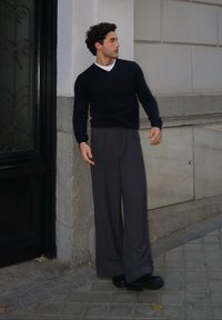 Granatowy sweter z dekoltem w serek, w zestawieniu z szarymi szerokimi spodniami i czarnymi butami. Gładka faktura, luźny krój, stojąc przed kamiennym murem.