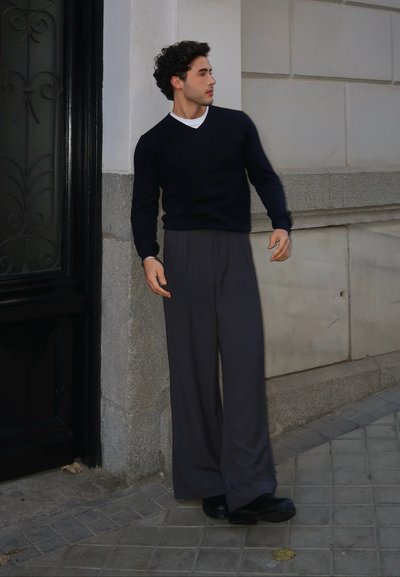 Jersey azul marino con cuello en V, combinado con pantalones de pierna ancha grises y zapatos negros. Textura suave, ajuste casual, de pie contra una pared de piedra.