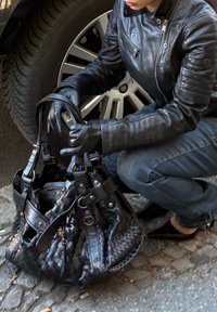 Schwarze Lederhandtasche mit geflochtenem Muster, mehreren Trägerschlaufen und verschiedenen Hardware-Akzenten. Auf Kopfsteinpflaster in der Nähe eines Fahrzeugreifens platziert.