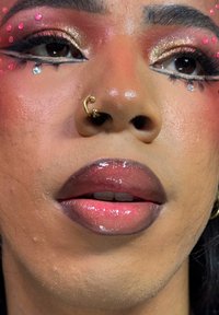 Primer plano de un rostro con labios brillantes con efecto degradado, delineador de ojos dorado y negro, pedrería rosa y plateada, y un aro dorado en la nariz.