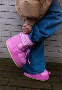Pink Moon Boot with a padded design, rounded shape, and contrasting white branding; it features laces and tassels, set against a brick background.