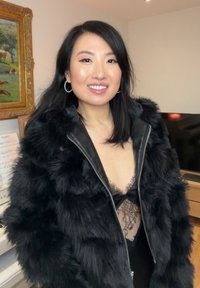 Femme souriante aux cheveux noirs portant des boucles d'oreilles anneaux, une veste noire en fourrure sur un haut en dentelle, debout à l'intérieur avec un tableau encadré et une télévision en arrière-plan.