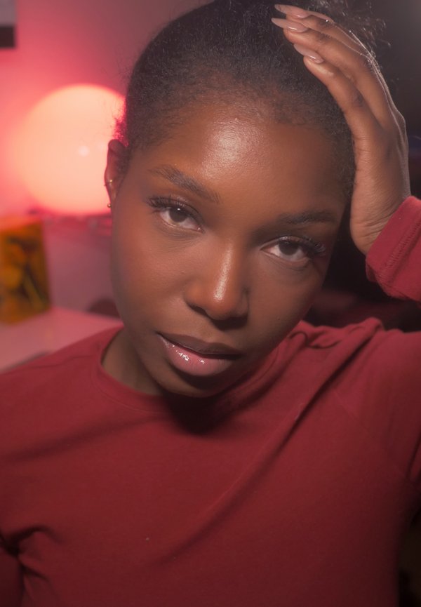 Primer plano de una persona con piel oscura y cabello recogido, que lleva una camiseta de manga larga color granate. Fondo con iluminación suave.