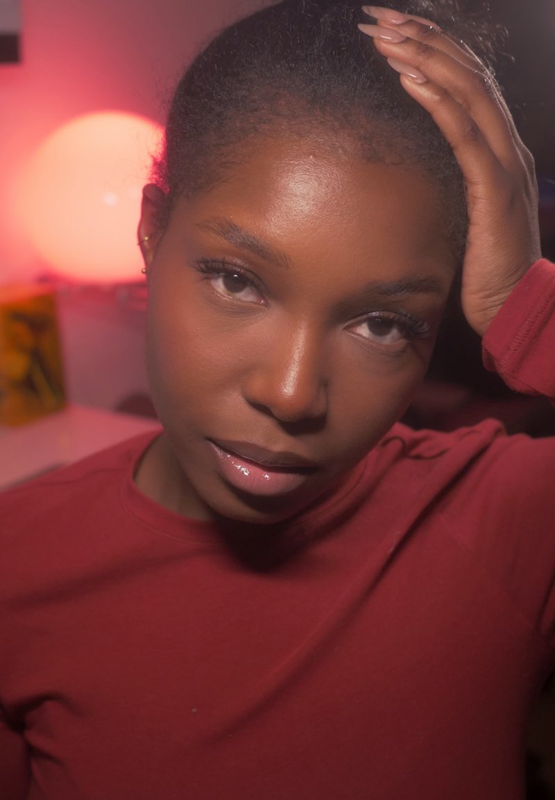 Primo piano di una persona che indossa una maglia a maniche lunghe color marsala. Ha la pelle scura, una texture liscia e i capelli raccolti. Illuminazione soffusa sullo sfondo.