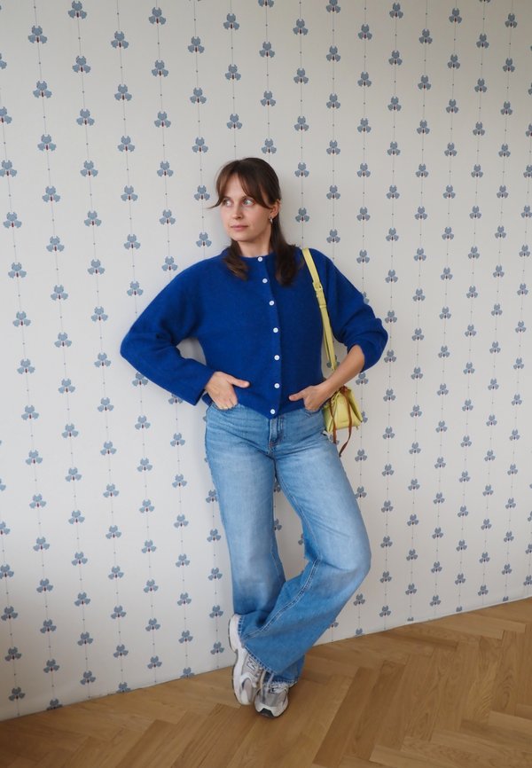 Mujer con cárdigan azul y vaqueros de pie junto a una pared con patrón, con las manos en los bolsillos, calzando zapatillas deportivas y llevando un bolso amarillo al hombro.