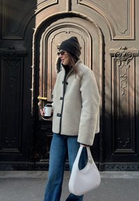 Casaco de fleece bege com botões pretos, gorro escuro, óculos de sol, jeans azuis e uma bolsa branca, segurando uma chávena de café. Porta de madeira detalhada ao fundo.