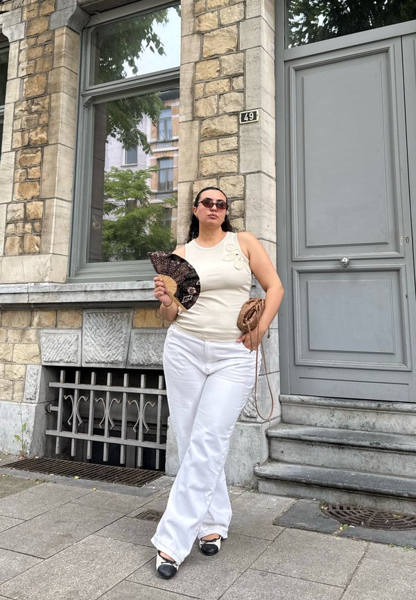 Femme portant des lunettes de soleil, un haut sans manches et un pantalon blanc, debout sur un trottoir en pierre tenant un éventail décoratif et un petit sac devant la porte d’un bâtiment.